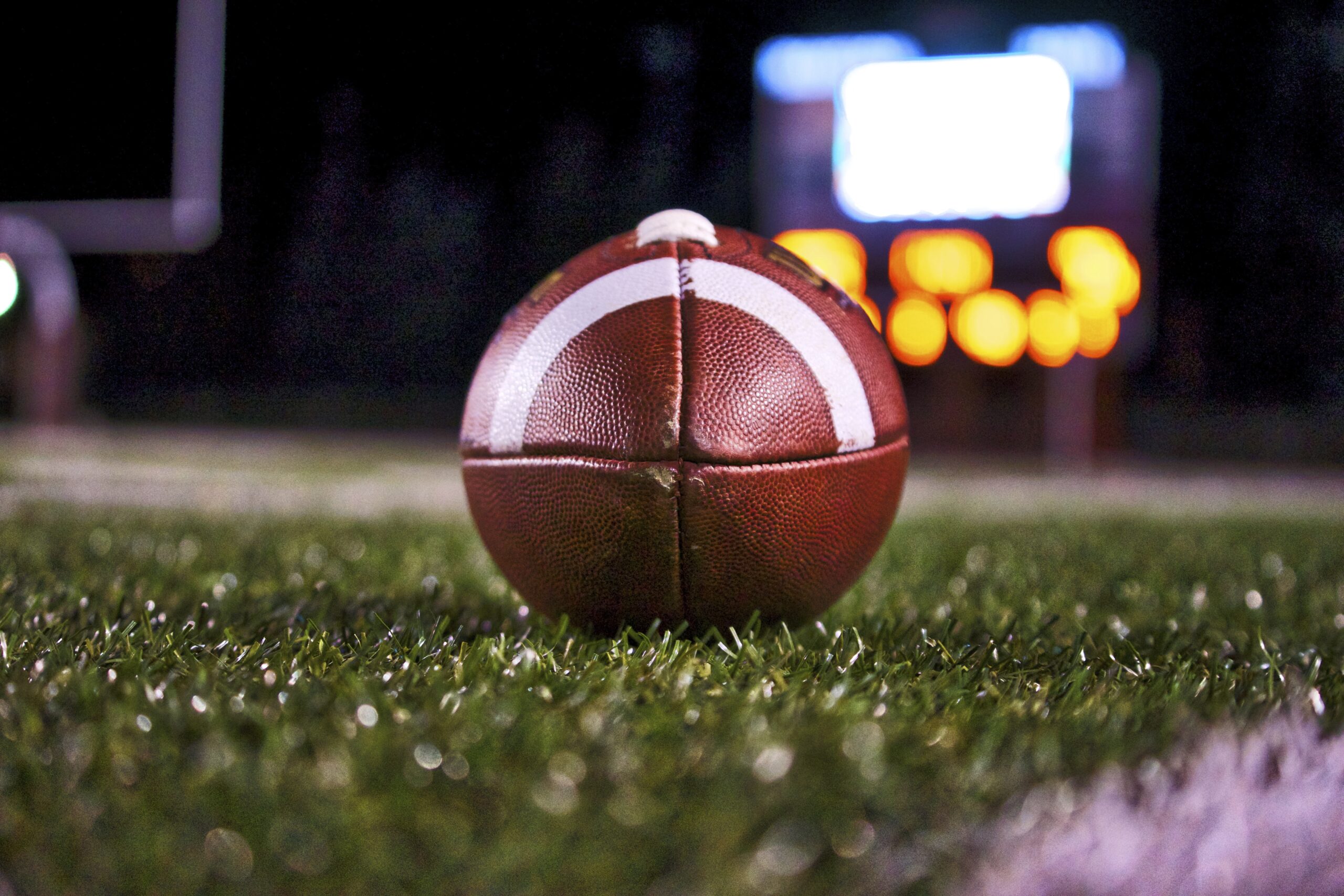 a football on a football field with a blurry scoreboard in the background.